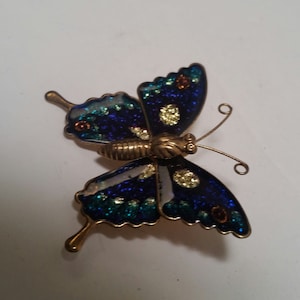May include: A blue and green butterfly brooch with gold accents. The butterfly has a glittery finish and is made of metal.