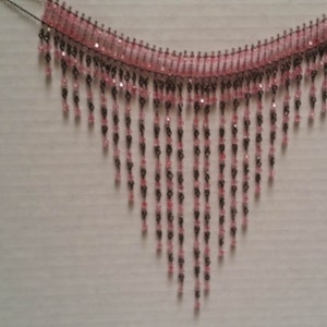 Pode incluir: Um colar de contas rosa e preto com um design de franjas. O colar &eacute; feito de pequenas contas que s&atilde;o enfiadas em um fio. A franja &eacute; feita de longos fios de contas que pendem do colar.