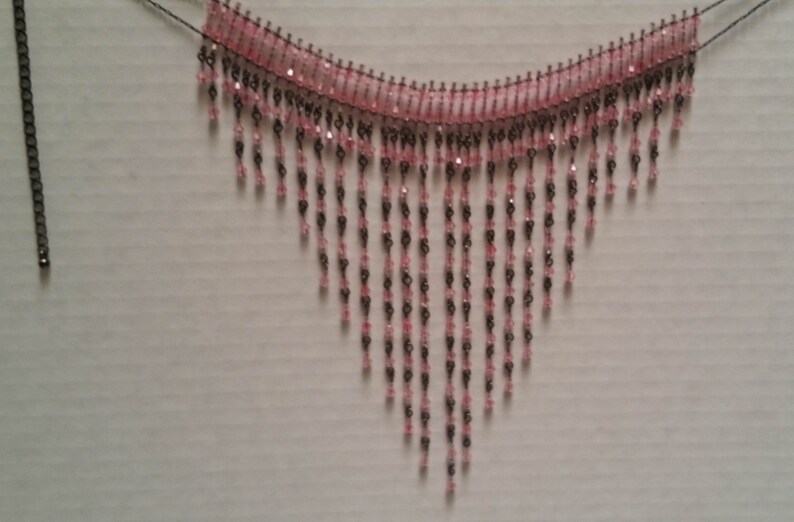 Pode incluir: Um colar de contas rosa e preto com um design de franjas. O colar &eacute; feito de pequenas contas que s&atilde;o enfiadas em um fio. A franja &eacute; feita de longos fios de contas que pendem do colar.