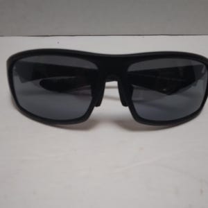 May include: Black sunglasses with dark gray lenses. The sunglasses have a thick, rectangular frame and a bridge over the nose. The lenses are reflective, and the sunglasses are set against a white background.