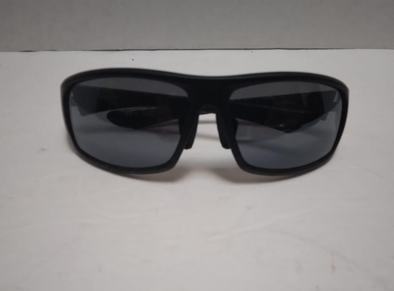 May include: Black sunglasses with dark gray lenses. The sunglasses have a thick, rectangular frame and a bridge over the nose. The lenses are reflective, and the sunglasses are set against a white background.