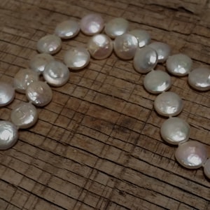 May include: A white pearl necklace with a silver clasp. The pearls are round and have a smooth, glossy finish. The necklace is laid out on a wooden surface.