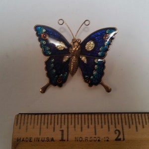 May include: A blue and gold butterfly brooch with a glittery finish. The butterfly is about 1.5 inches wide.