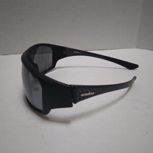 May include: Black Ironman sunglasses with a wraparound design. The frames are black with a carbon fiber pattern on the arms. The lenses are dark, providing protection from the sun. The Ironman logo is visible on the arm.