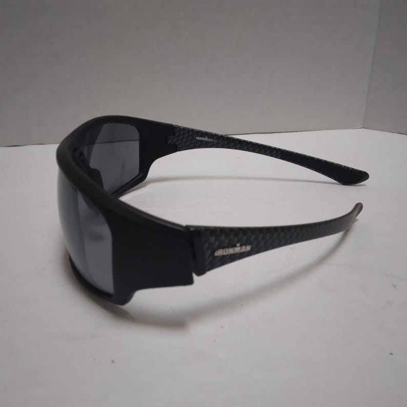 May include: Black Ironman sunglasses with a wraparound design. The frames are black with a carbon fiber pattern on the arms. The lenses are dark, providing protection from the sun. The Ironman logo is visible on the arm.