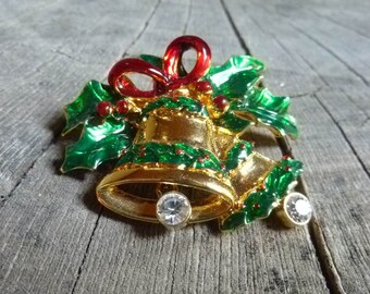 May include: A gold-colored Christmas bell brooch with a red bow and green holly leaves. The brooch features two clear, round gemstones and small red berry accents, perfect for holiday attire.