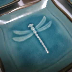 May include: Square teal ceramic plate with a raised dragonfly design in the center. The dragonfly is white and has delicate wings. The plate has a brown rim and a slightly textured surface.