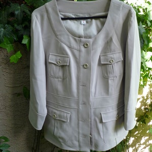 May include: A light beige jacket with a rounded collar and four front pockets. The jacket has three buttons down the front and three-quarter length sleeves. It is hanging on a black hanger against a background of green foliage.