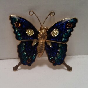 May include: A gold-toned butterfly brooch with blue, green, and yellow enamel accents. The butterfly has a glittery finish.