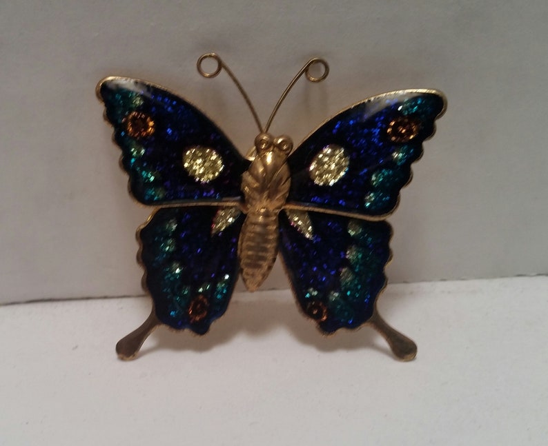 May include: A gold-toned butterfly brooch with blue, green, and yellow enamel accents. The butterfly has a glittery finish.