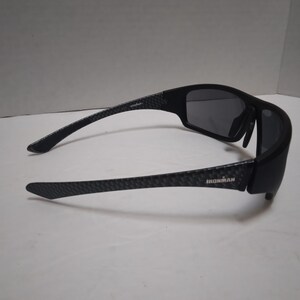 May include: Black sunglasses with dark gray lenses. The arms have a textured pattern and the word "IRONMAN" is printed on the side. The sunglasses are designed for sports and outdoor activities.