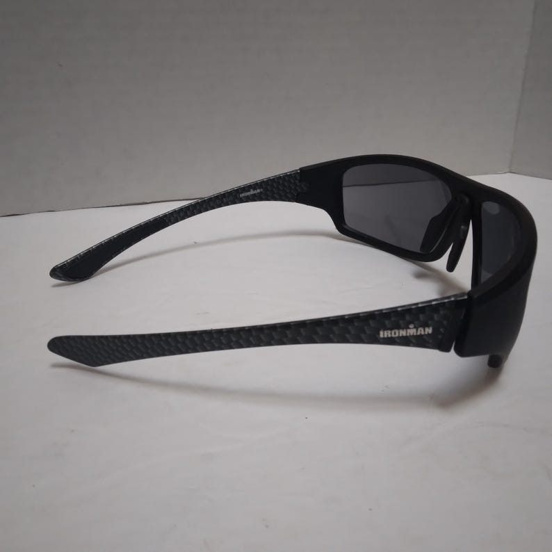 May include: Black sunglasses with dark gray lenses. The arms have a textured pattern and the word "IRONMAN" is printed on the side. The sunglasses are designed for sports and outdoor activities.
