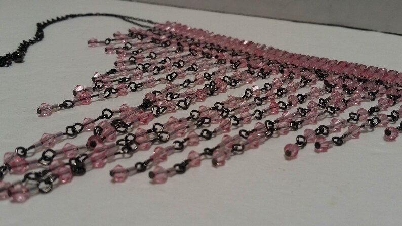 Pode incluir: Um colar de contas rosa e preto com um design de franja. O colar &eacute; feito de pequenas contas rosa enfiadas em uma corrente preta.