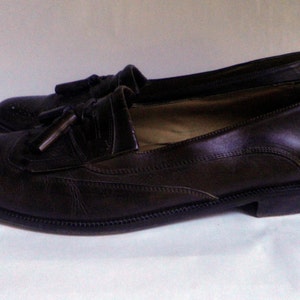Puede incluir: Un par de mocasines marrones de cuero con borlas y dise&ntilde;o brogue. Los zapatos tienen punta puntiaguda y tac&oacute;n bajo.