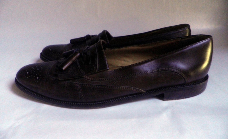 Puede incluir: Un par de mocasines marrones de cuero con borlas y dise&ntilde;o brogue. Los zapatos tienen punta puntiaguda y tac&oacute;n bajo.