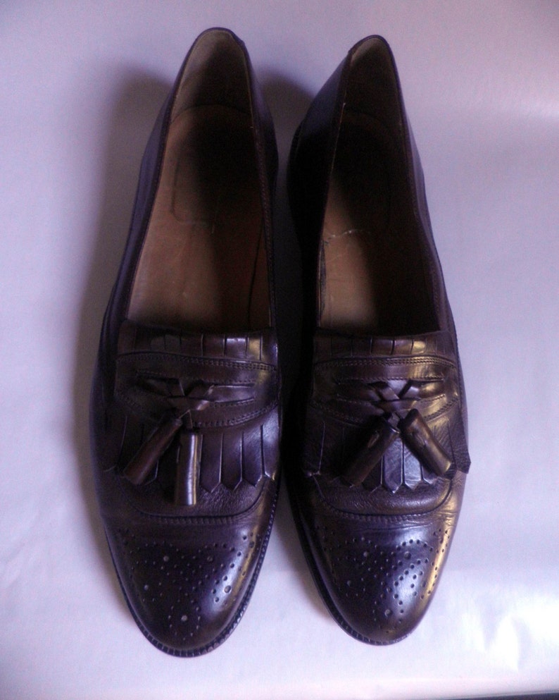 Puede incluir: Un par de mocasines marrones de cuero con borlas y dise&ntilde;o brogue. Los zapatos tienen una punta puntiaguda y un detalle de borlas en el empeine.