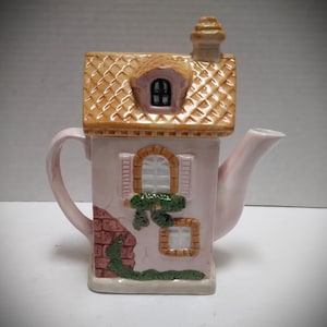 May include: A ceramic teapot shaped like a house, featuring a light pink body with a tan roof and chimney. The teapot has a handle, spout, and decorative brick and window details. Green vines and a small window are also visible.