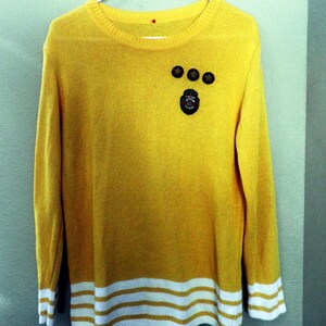 May include: A yellow long-sleeved sweater with white stripes at the bottom. The sweater has three small round buttons and a small patch with a design on the front.