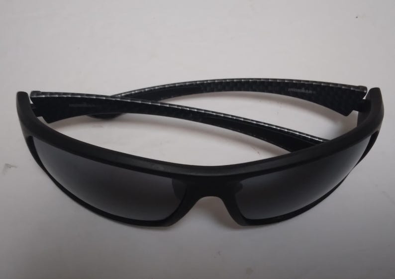 May include: Black sunglasses with a matte finish and dark lenses. The arms have a textured pattern. These sunglasses are designed for sports and outdoor activities.