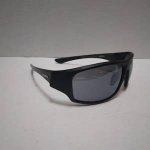 May include: Black sunglasses with mirrored lenses. The sunglasses have a thick, rectangular frame and a curved design. The brand name "IRONMAN" is visible on the left arm. The lenses are a reflective silver color.