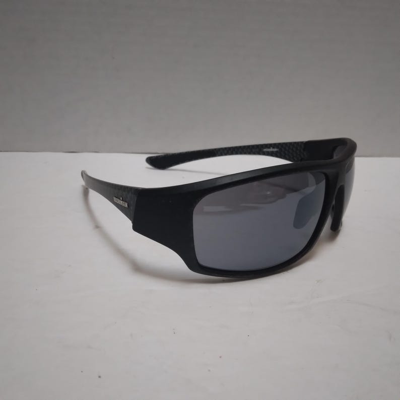 May include: Black sunglasses with mirrored lenses. The sunglasses have a thick, rectangular frame and a curved design. The brand name "IRONMAN" is visible on the left arm. The lenses are a reflective silver color.