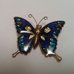 May include: A blue and green butterfly brooch with gold accents. The butterfly has glitter on its wings.