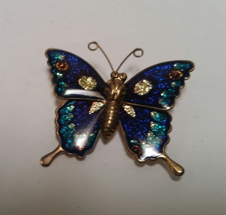 May include: A blue and green butterfly brooch with gold accents. The butterfly has glitter on its wings.