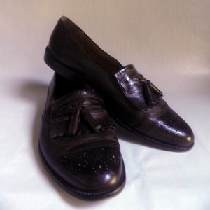 Puede incluir: Un par de mocasines marrones de cuero con borlas y detalles de brogue. Los mocasines tienen una punta puntiaguda y una borla en el empeine.