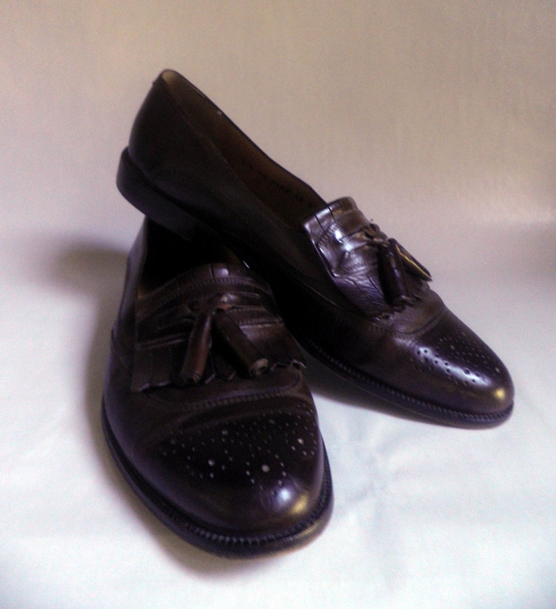 Puede incluir: Un par de mocasines marrones de cuero con borlas y detalles de brogue. Los mocasines tienen una punta puntiaguda y una borla en el empeine.