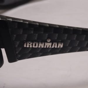 May include: Close-up of a black and gray patterned sunglasses arm. The text "IRONMAN" is displayed in silver lettering. The arm has a textured, checkered design.