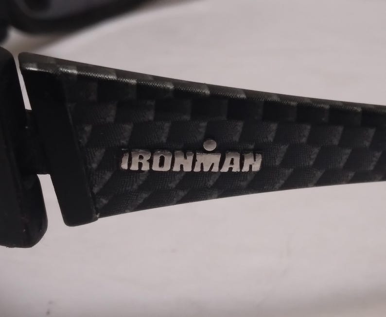May include: Close-up of a black and gray patterned sunglasses arm. The text "IRONMAN" is displayed in silver lettering. The arm has a textured, checkered design.