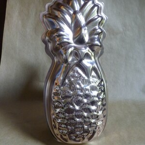 May include: A silver pineapple-shaped cake mold with a detailed design. The mold has a loop at the top for hanging.