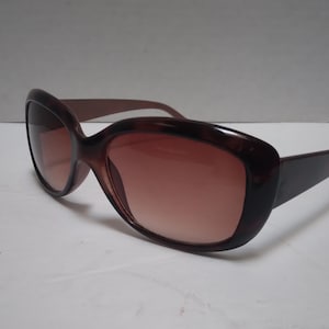May include: Brown oversized sunglasses with a gradient lens. The frames are a dark brown color, and the lenses transition from a darker shade at the top to a lighter shade at the bottom. The sunglasses have a classic, rounded shape.