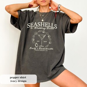 May include: A pepper-coloured t-shirt with an ivory design. The shirt displays the text "SEASHELLS & Wedding Bells" above a martini glass, ring, and seashells graphic. Below is "Emily's Bachelorette, Napa Valley, CA" and the year "2026".