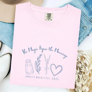May include: A light pink Comfort Colors t-shirt with the text "The Magic Before the Marriage" and a graphic of a salt shaker, herbs, lavender, and a heart. The text "EMILY'S BACH / EST. 2025" is also printed on the shirt.