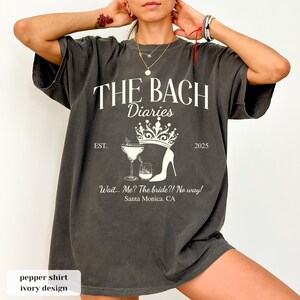 May include: Pepper-colored t-shirt with the text "THE BACH Diaries" in white. The design includes a crown, high heel, and cocktail glasses. The text "Wait... Me? The bride?! No way! Santa Monica, CA" is also printed on the shirt.