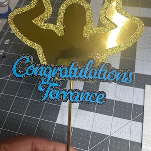 May include: A gold glitter cake topper featuring a silhouette of a person with arms raised in celebration. The words "Congratulations Terrance" are written in blue script with a black glitter outline. The topper is attached to a gold stick.