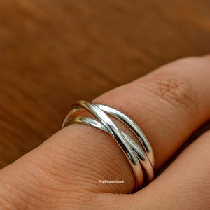 以下が含まれることがあります： 3つのバンドが絡み合ったシルバーカラーのリング。バンドは滑らかで磨かれており、光を反射しています。指にはめられており、背景はぼやけた木目です。