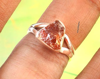 May include: A silver ring featuring a raw, reddish-brown gemstone. The stone has a rough, natural texture and is set in a split-shank band. The ring is displayed on a finger against a blurred, yellow-green background.