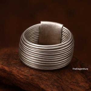 May include: A silver ring with a wide, textured band composed of parallel lines. The ring includes a rectangular accent. It is presented on a dark brown wooden surface. TheElegantAura is printed on the bottom right.