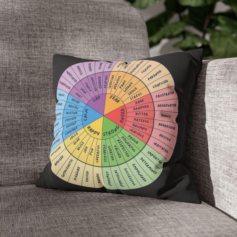 Puede incluir: Coj&iacute;n cuadrado negro con un dise&ntilde;o de rueda de emociones colorida. La rueda est&aacute; dividida en secciones etiquetadas con varios sentimientos como feliz, triste y enojado. El coj&iacute;n est&aacute; sobre un sof&aacute; gris.