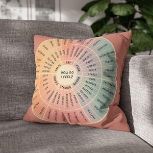 May include: A square throw pillow in a muted coral shade featuring a circular emotion wheel graphic. The center text reads "HOW DO I FEEL?" surrounded by radiating segments labeled with various emotions in black text, ranging from calm to anger.