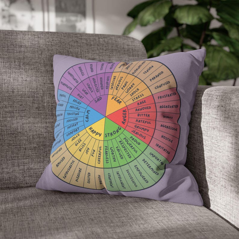 Puede incluir: Un coj&iacute;n cuadrado lavanda con un dise&ntilde;o de rueda de emociones colorida. La rueda est&aacute; dividida en secciones etiquetadas con varios sentimientos como "Feliz", "Triste" y "Miedo". El coj&iacute;n est&aacute; sobre un sof&aacute; gris.