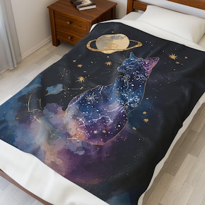 May include: A dark blue blanket featuring a cat silhouette filled with a galaxy design in shades of blue, purple, and gold. The blanket also includes a planet with rings and gold stars, creating a celestial theme.