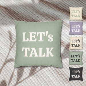 May include: A sage green square pillow with the text "LET'S TALK" in white block letters. The image also shows five smaller pillows in various colors, each with the same text. The pillows are on a textured, neutral-toned surface.