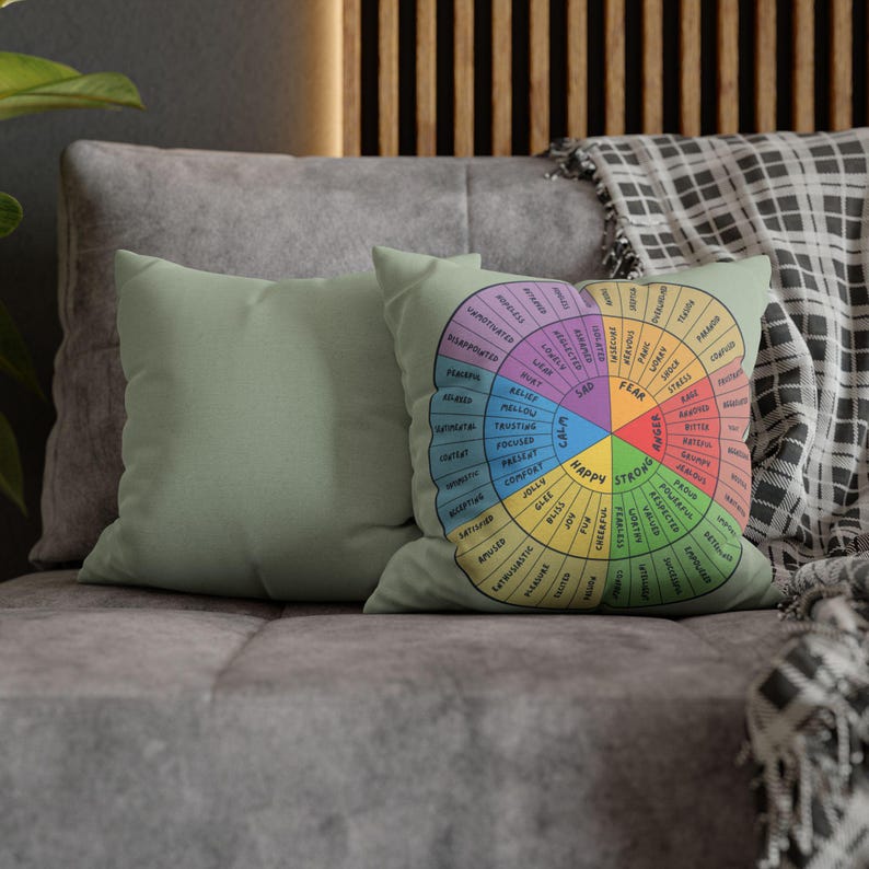 Puede incluir: Dos cojines decorativos sobre un sof&aacute; gris. Uno es verde claro liso, el otro presenta un dise&ntilde;o de rueda colorida con palabras como "feliz", "triste" y "miedo". El dise&ntilde;o es una representaci&oacute;n visual de las emociones.