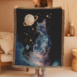 May include: A woven throw blanket featuring a celestial cat design. The cat is silhouetted against a dark blue background with a galaxy design. A planet with a ring and gold stars are also featured. The blanket has a fringed edge.