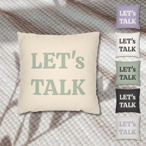 May include: Cream-colored square pillow with the text "LET'S TALK" in a soft green hue. The image also shows smaller pillows in various colors, each with the same text. The pillows are arranged on a textured, off-white surface.