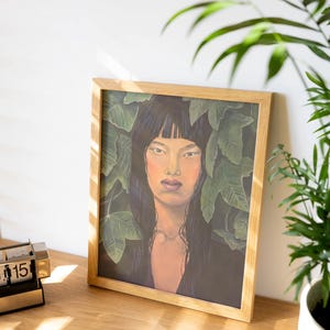 Könnte beinhalten: Ein gerahmter Kunstdruck mit einem Porträt einer Frau mit langen schwarzen Haaren und neutralem Gesichtsausdruck, umgeben von grünen Blättern. Das Kunstwerk befindet sich in einem hellen Holzrahmen, auf einer Holzoberfläche, mit einem kleinen Schreibtischkalender und einer Topfpflanze.