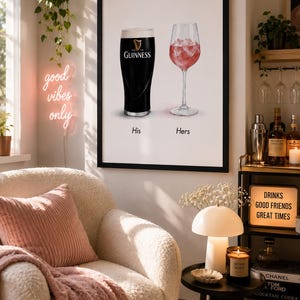 May include: A framed print featuring a pint of Guinness and a glass of ros&eacute;, labeled "His" and "Hers." The print hangs above a cozy armchair with a pink pillow and blanket. A neon sign reads "good vibes only."